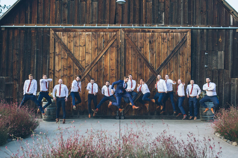 groomsmen and groom jumping in front of tin roof barn wedding venue